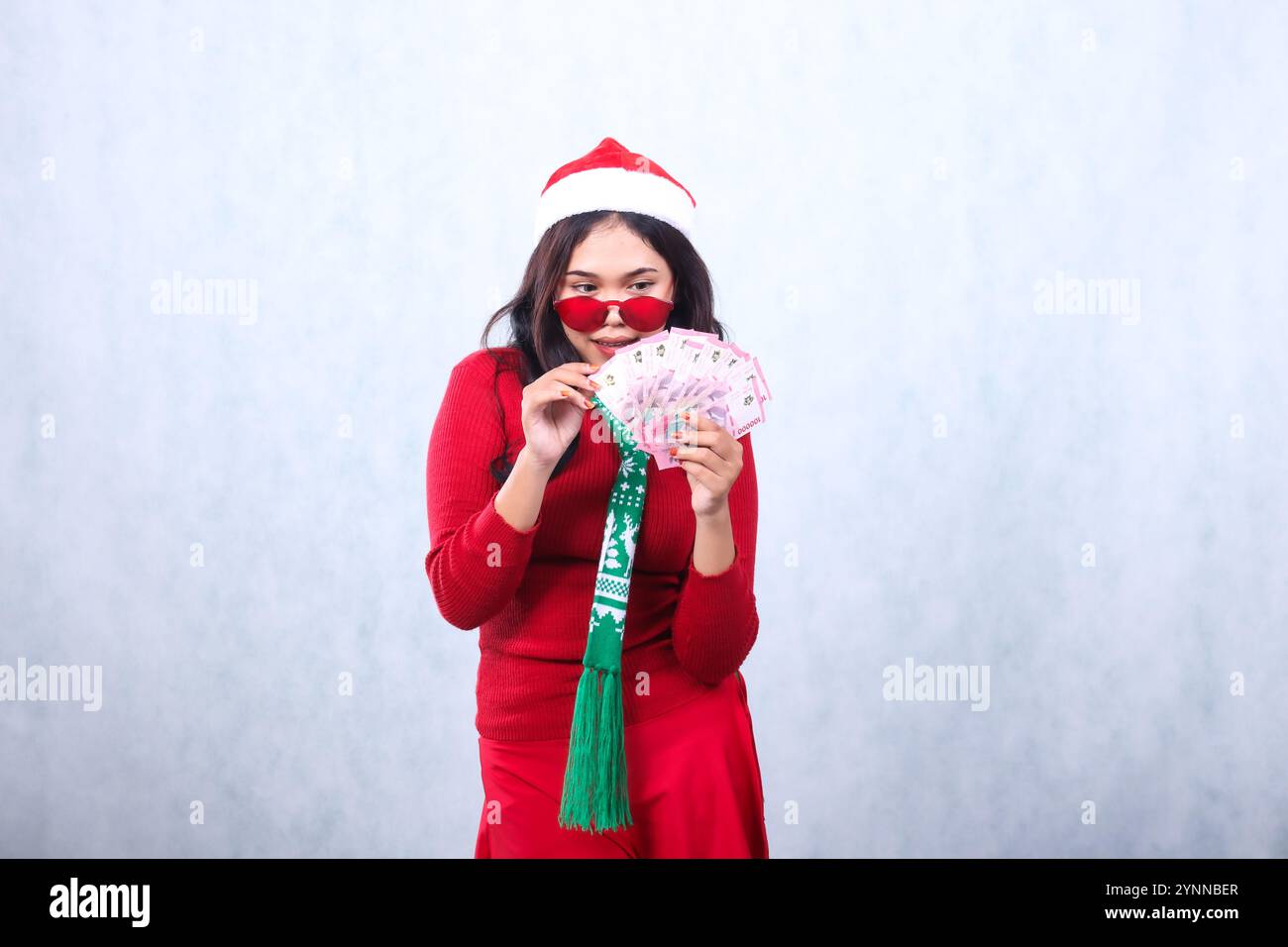 Charming adult woman wearing red christmas sweater, red glasses, Santa hat and scarf, focus counting money, hand holding cash rupiah, isolated on whit Stock Photo