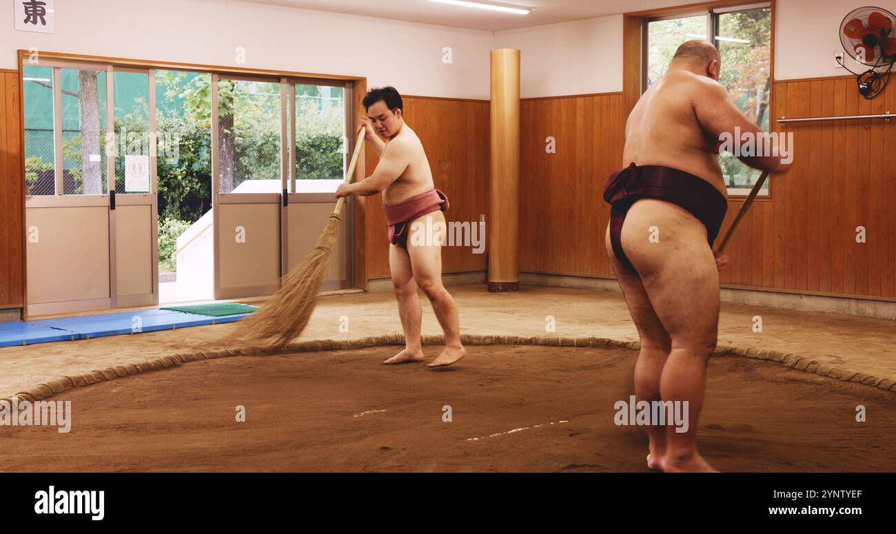 Ceremony, broom and dohyo with Sumo wrestler people sweeping sand in mawashi, getting ready for competition. Cleaning, prepare and start with men in Stock Photo