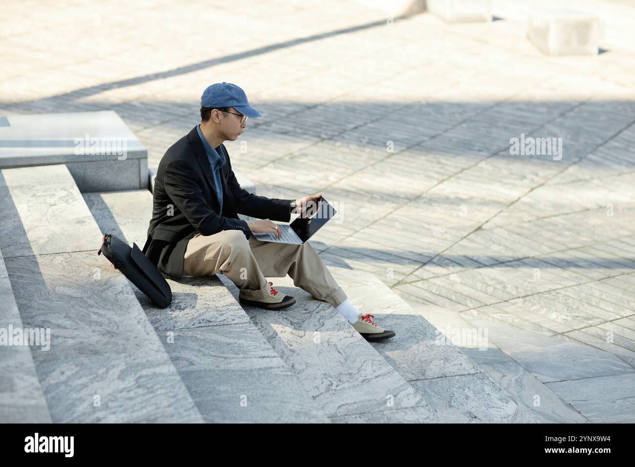Side view of Asian male IT developer typing codes on laptop working outside in sunlight while sitting on concrete steps against paved square in city s Stock Photo