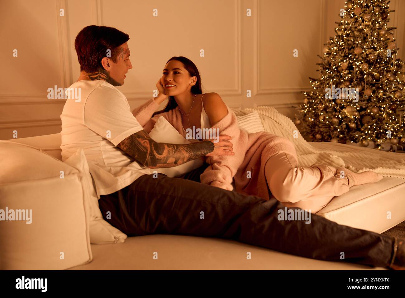 A couple embraces warmly on a couch surrounded by soft lights and a decorated Christmas tree. Stock Photo