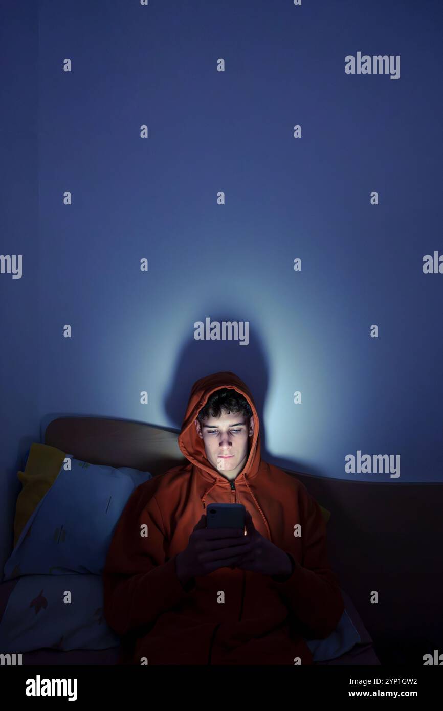 Teenager using mobile phone sitting on bed at night, illuminated by screen light Stock Photo