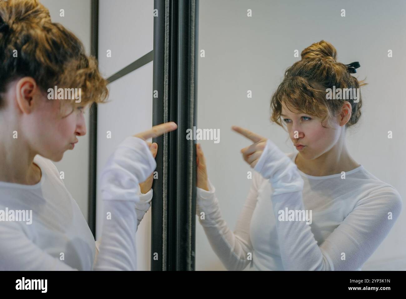 Young woman with curly hair tied in a bun, wearing a white long-sleeved shirt, points at her reflection in a mirror with a displeased expression Stock Photo
