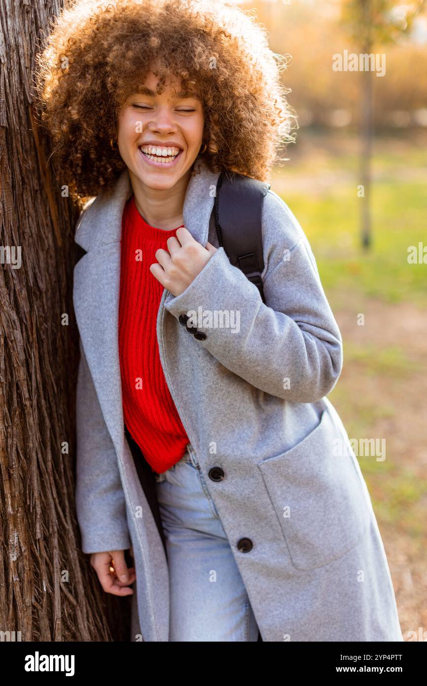 Smiling young woman with curly hair stands against a tree, wearing a ...