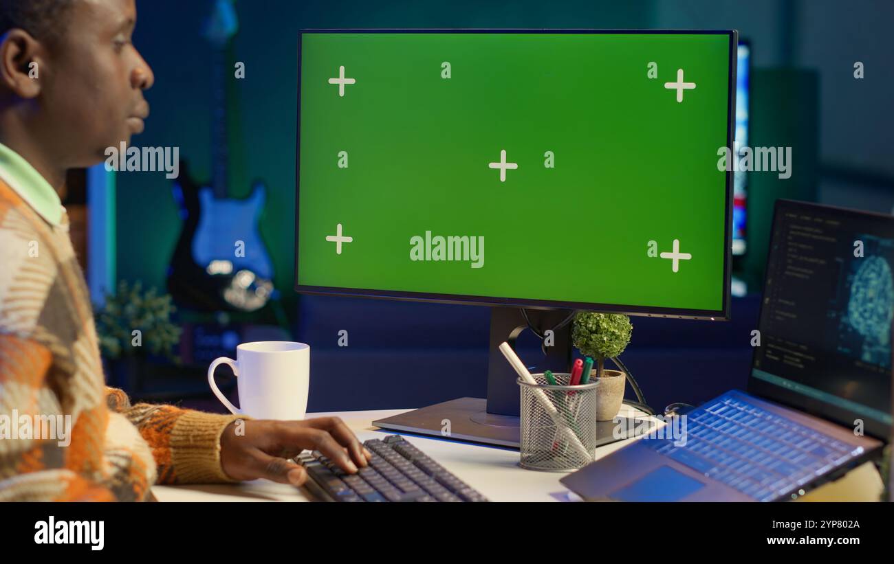 Black person programming neural network code with a green screen on computer to build databases. System engineer working from home while looking at data center security servers. Camera A. Stock Photo