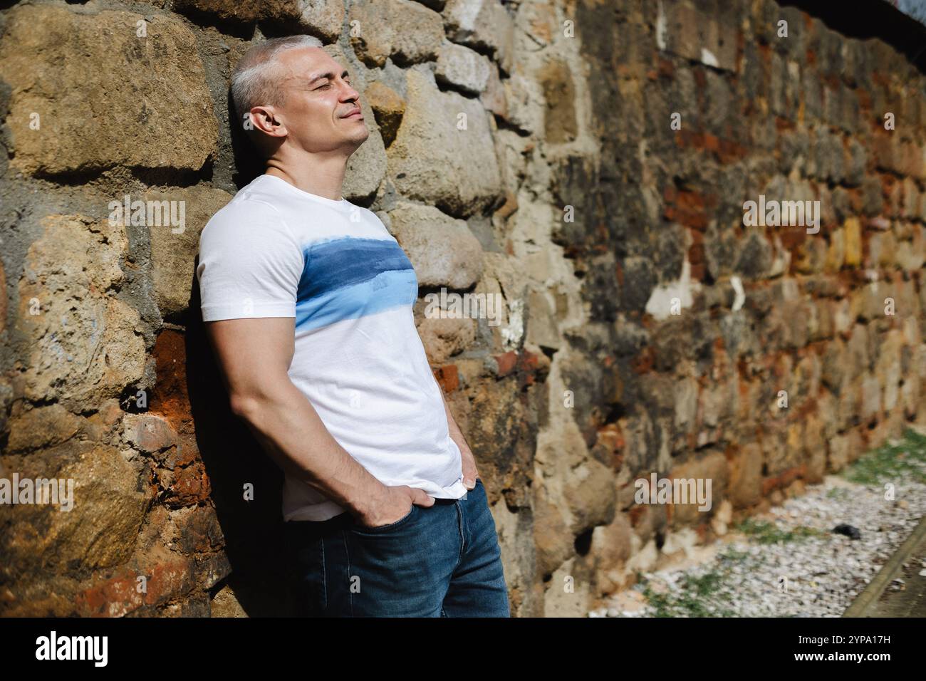 A man in a white shirt and blue jeans is leaning against a stone wall Stock Photo