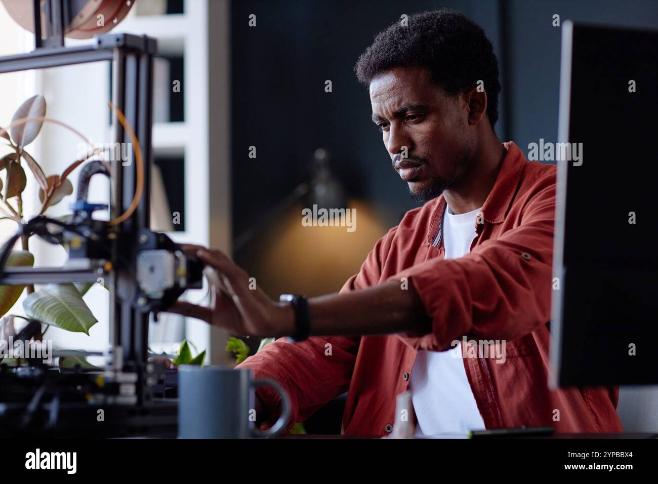 Man performing intricate work with a 3D printer, highlighting concentration and precision. The workspace includes plants and various tools emphasizing a creative environment Stock Photo