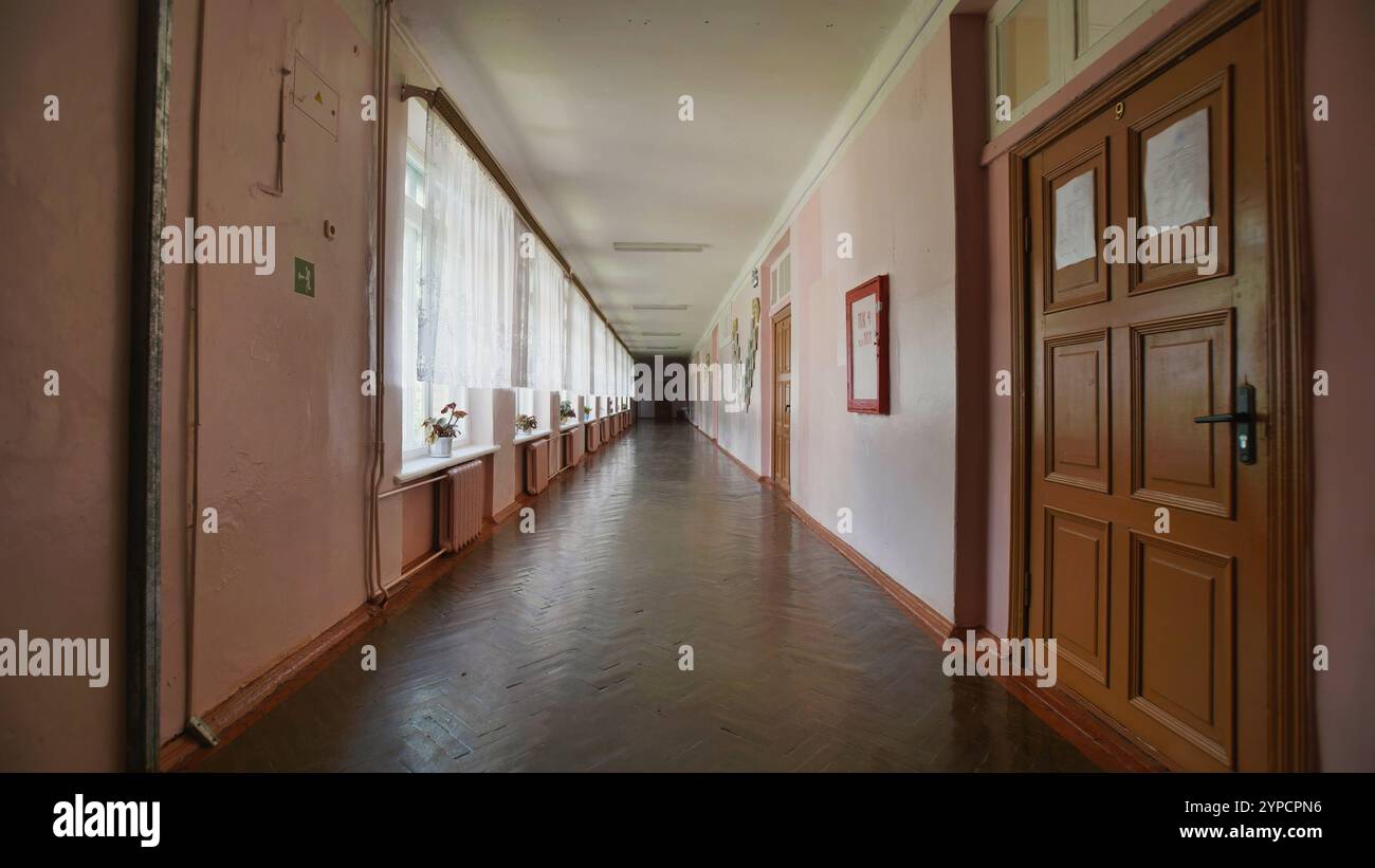 Deserted school hallway with pink walls, wooden doors, and windows adorned with curtains, evoking a sense of quietude on the last day of school Stock Photo