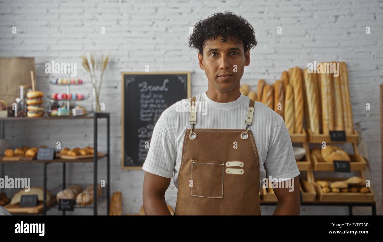 Hispanic man working in an indoor bakery, surrounded by fresh bread and pastries, wearing a white shirt and brown apron, with a neat interior showcasi Stock Photo
