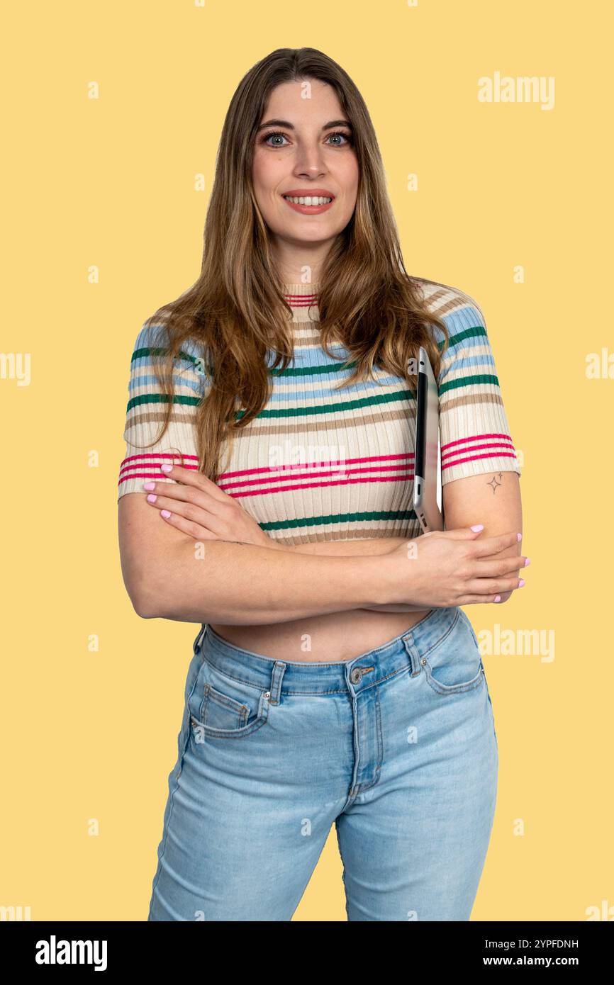 Portrait of a cheerful young woman holding a tablet under her arm, exuding confidence and approachability against a vibrant yellow backdrop Stock Photo