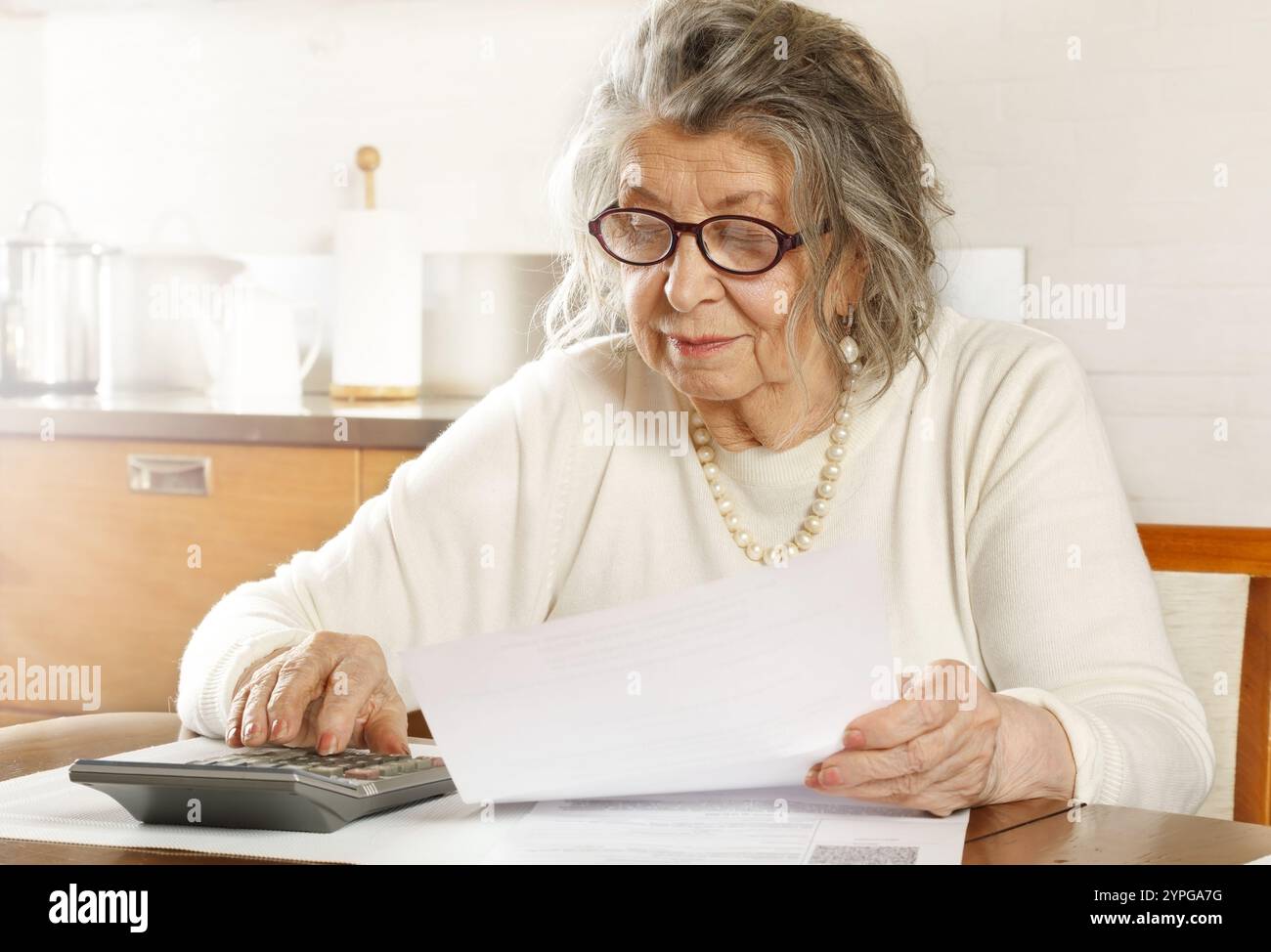 An old woman sitting at a table in the kitchen looking through receipts with a calculator Stock Photo