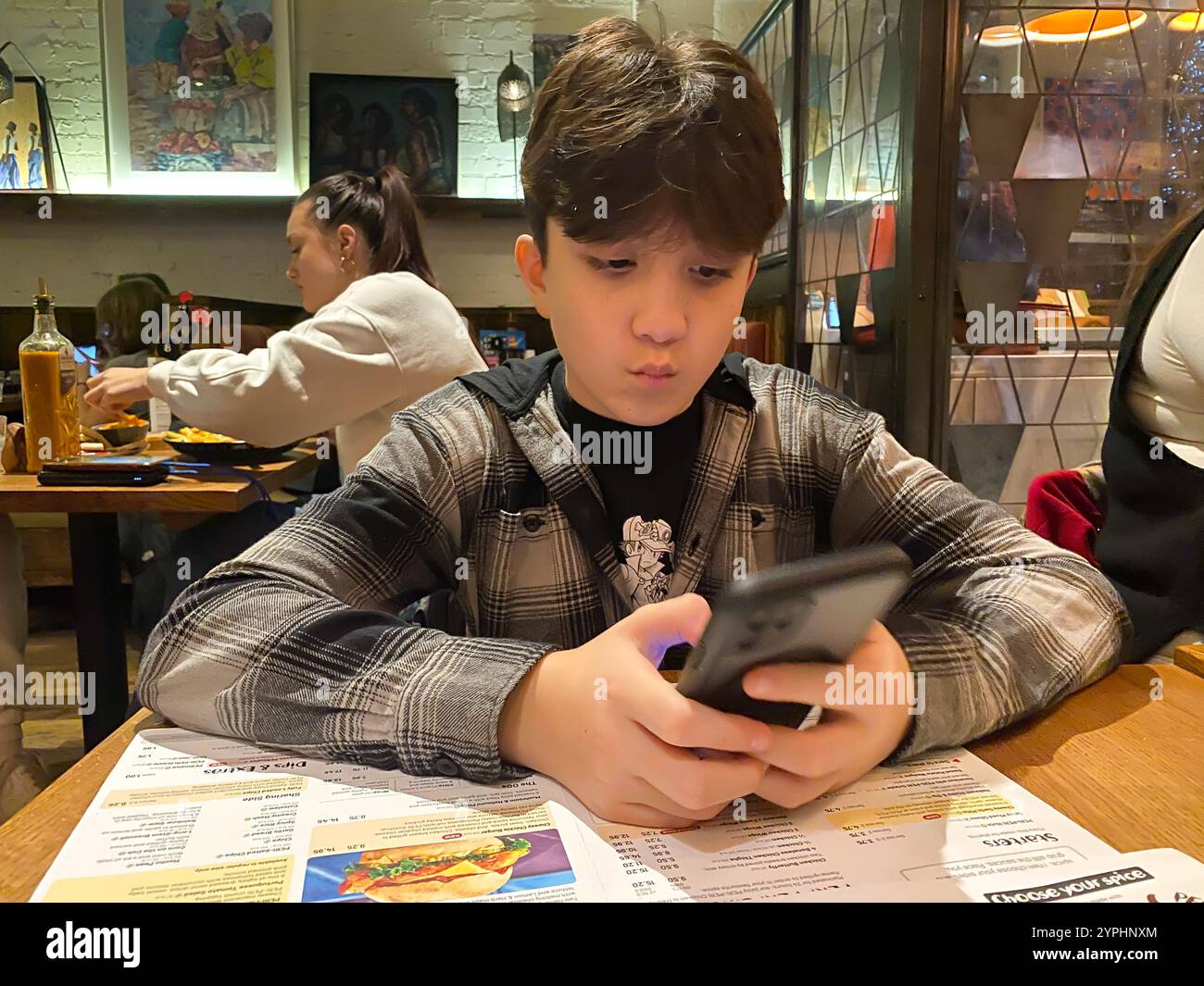 A boy sits at a table in a restaurant playing with his mobile phone. - Smartphone Captured Stock Image