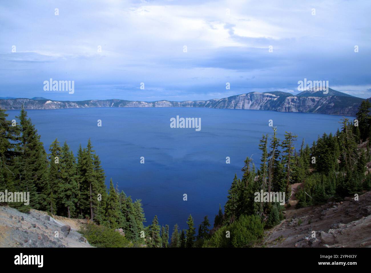 View across Crater Lake to the far lip of the crater rising in the background, sunlight sparkling on the pure blue water surface with tiny clouds. Stock Photo