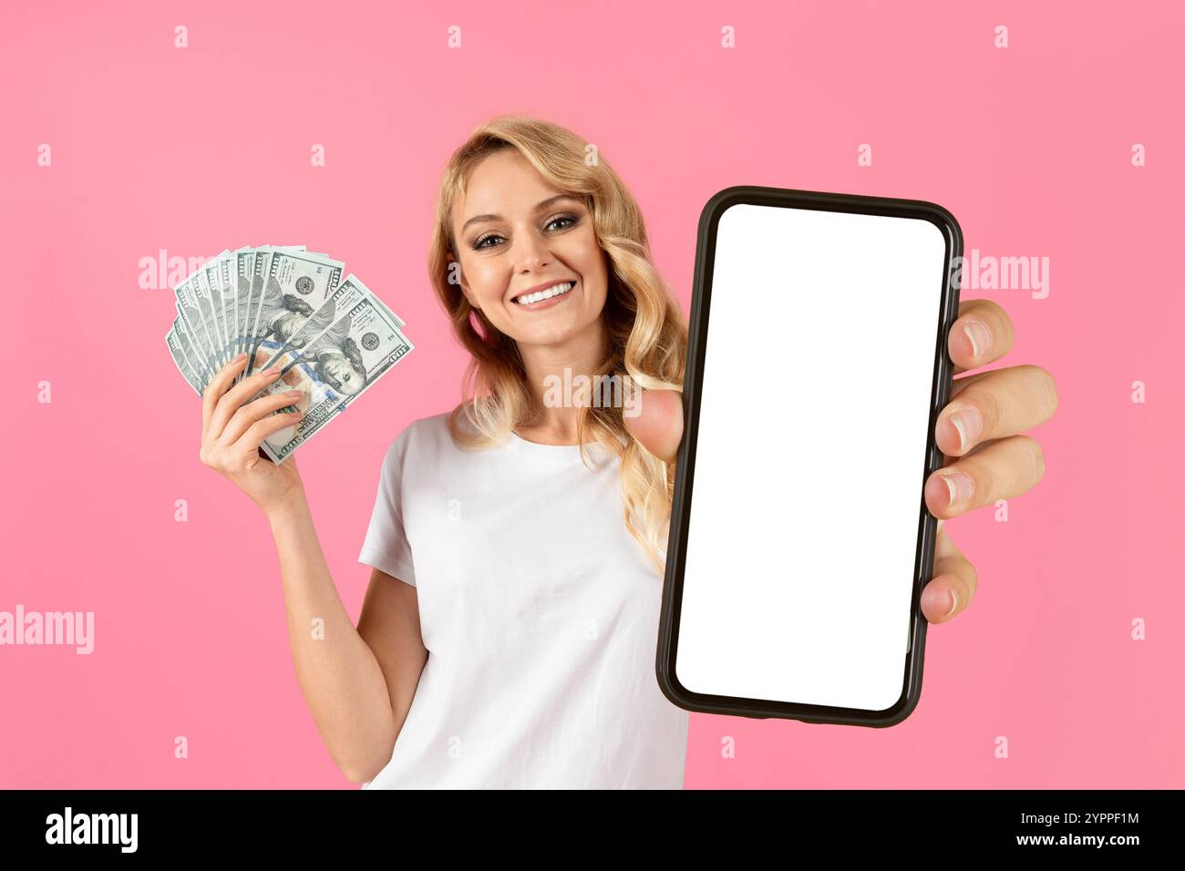 Young woman smiles while holding cash and a blank phone against pink backdrop Stock Photo