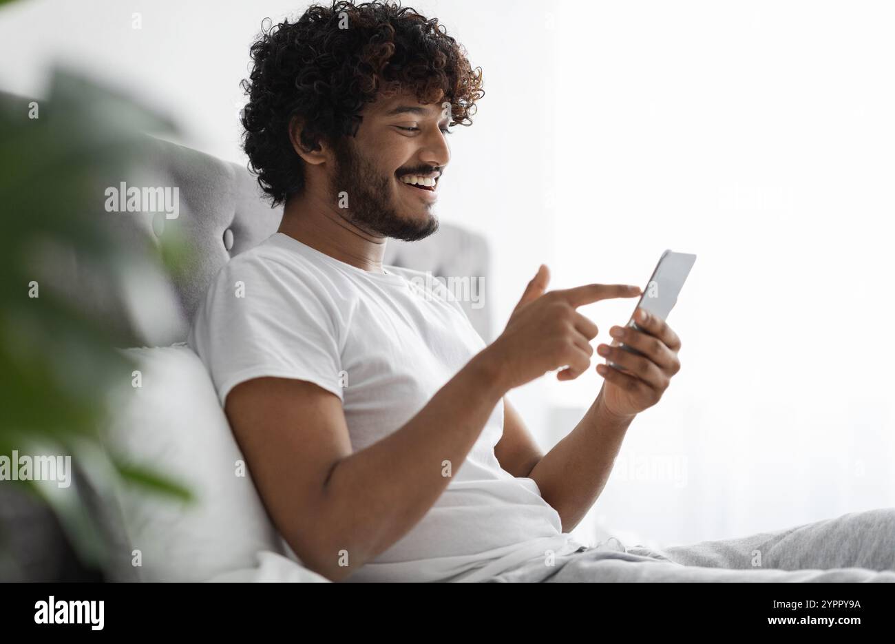 Positive dark-skinned guy scrolling in bed, using smartphone Stock Photo