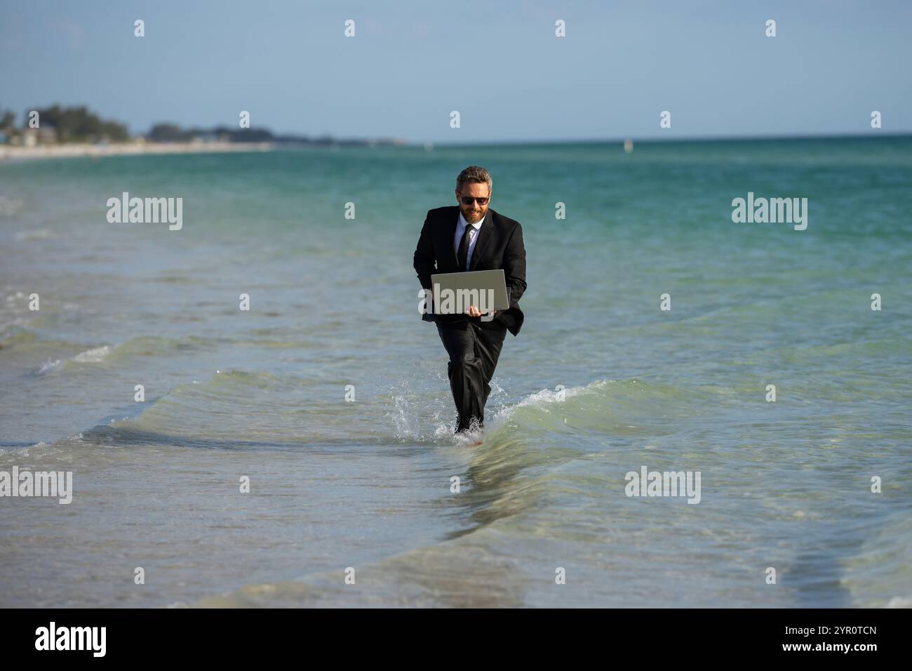 Business man in suit with laptop running in sea water. Travel tourism and business concept. Crazy male office employee with laptop running in sea Stock Photo