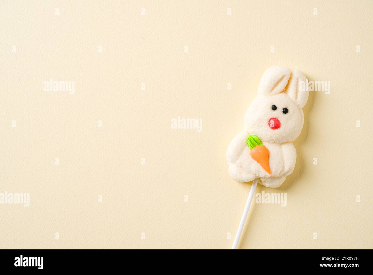 A cute bunny shaped marshmallow lollipop holding an orange carrot, set on a minimalist pastel background Stock Photo