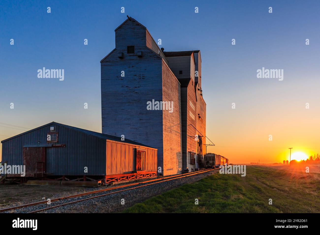 A train is pulling a large grain silo. The sun is setting in the background. The silo is old and the train is moving Stock Photo