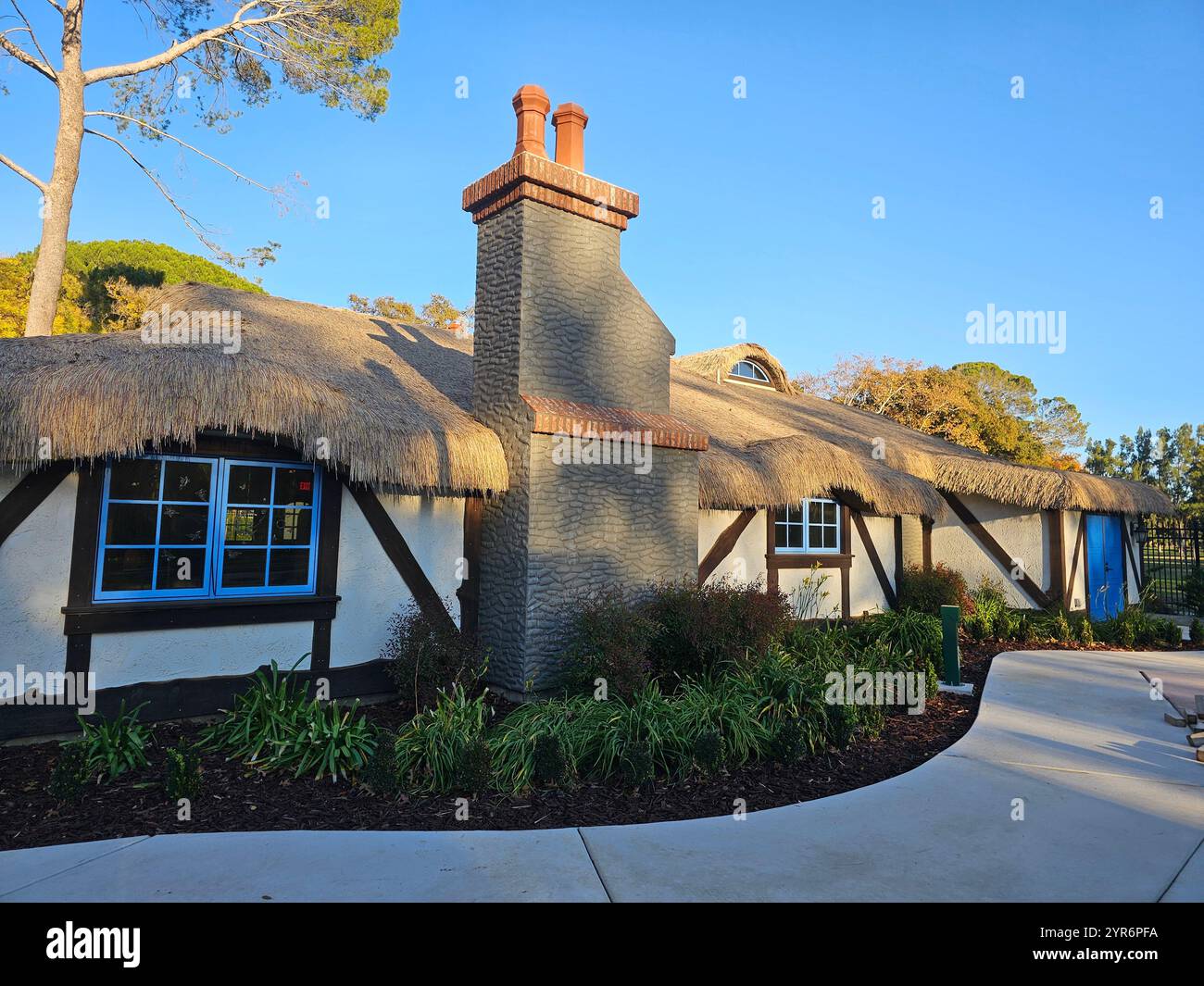 A charming thatched-roof cottage with rustic timber framing and bright blue windows, surrounded by greenery on a clear sunny day. - Smartphone Captured Stock Image