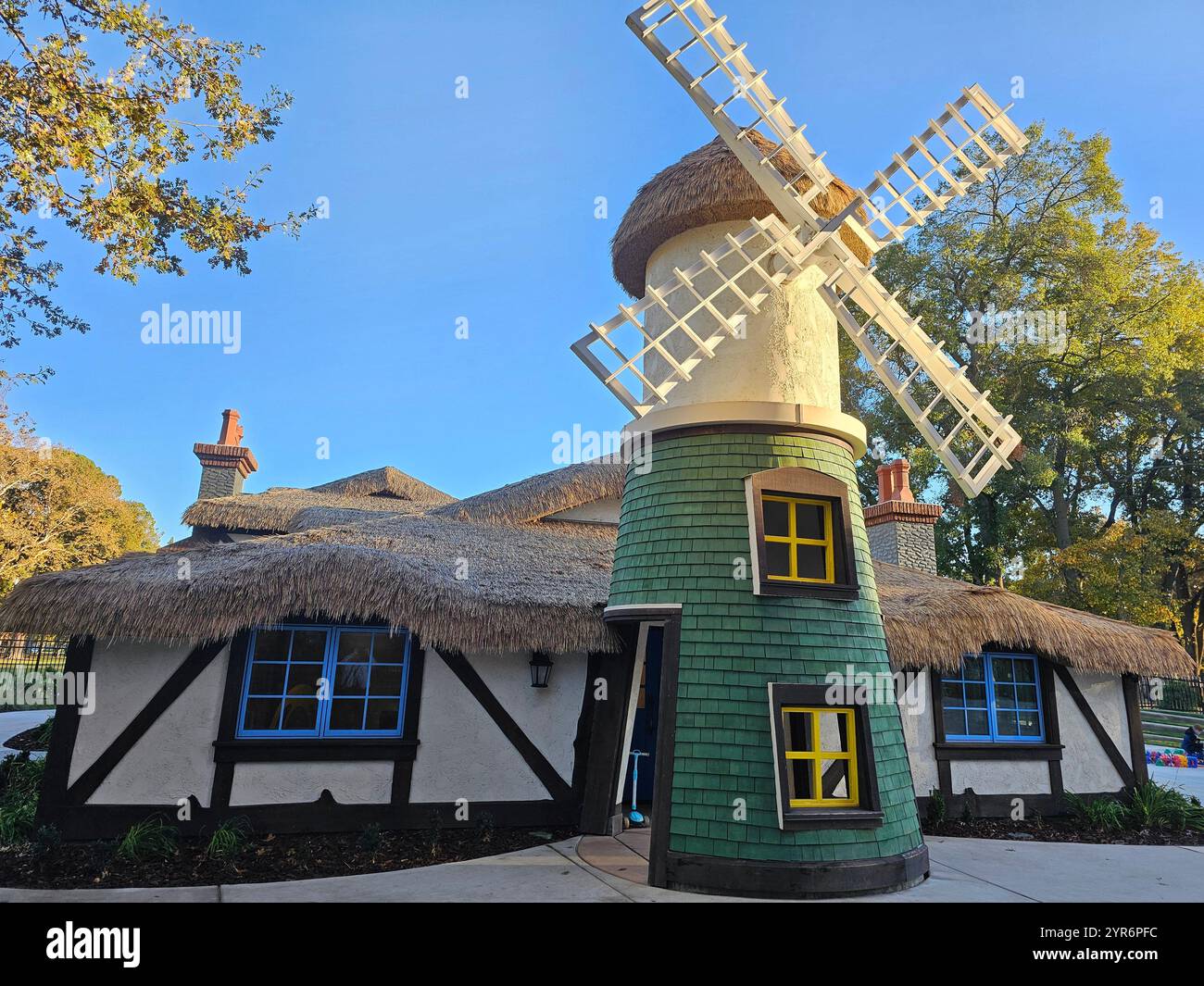 A charming windmill with a thatched roof and green shingles stands in a picturesque setting surrounded by trees on a clear, sunny day. - Smartphone Captured Stock Image