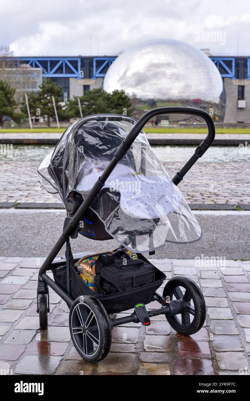 Baby sleeping in a stroller on a rainy day, protected by a rain cover with 'La Géode' dome in the background, along the Canal de l'Ourcq in Parc de la Stock Photo