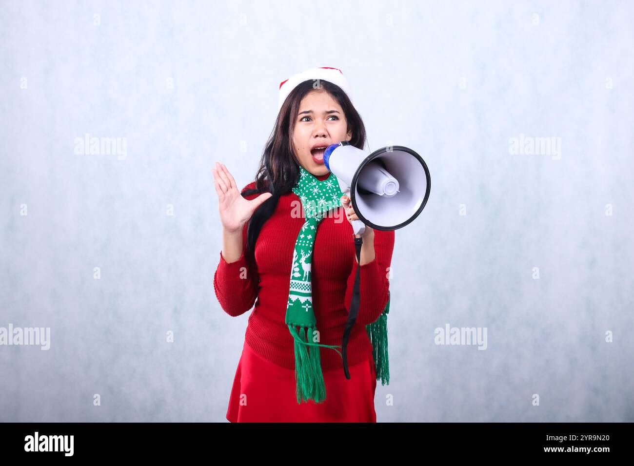 beautiful young asian woman wearing red christmas sweater with santa hat and scarf, screaming scared candid over holding megaphone loudspeaker and foc Stock Photo