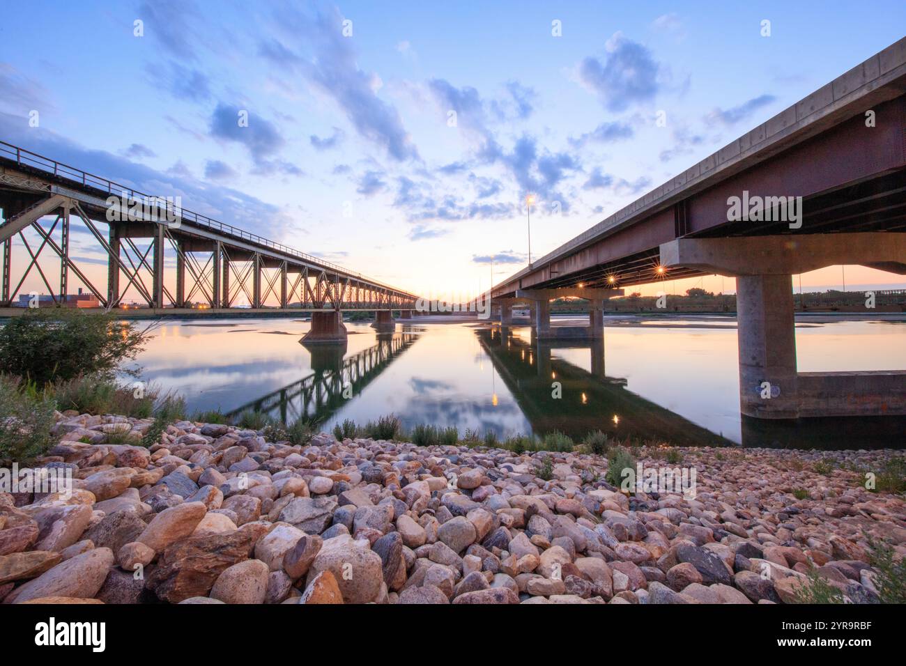 A bridge spans a river with a beautiful sunset in the background. The bridge is made of concrete and has a reflective surface Stock Photo