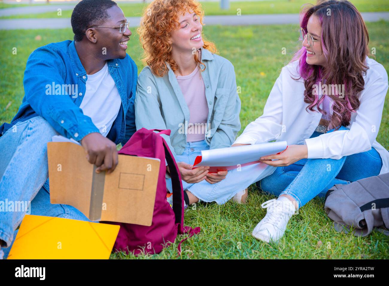 Group of diverse students sitting on grass and smiling while holding notebooks Stock Photo