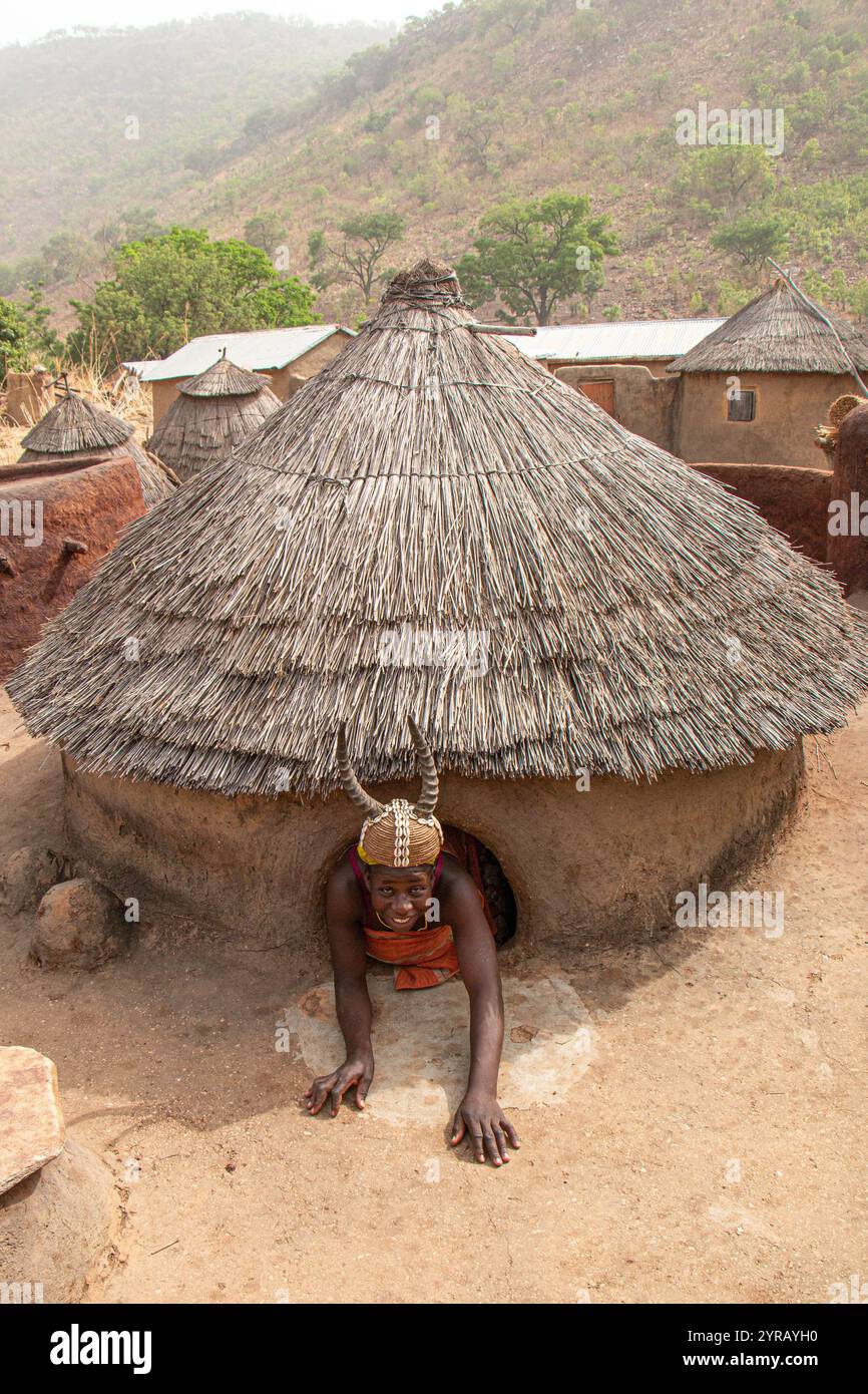 Woman in Traditional Costume with Horned Hat Emerging from a Clay House in a Togo Village Stock Photo