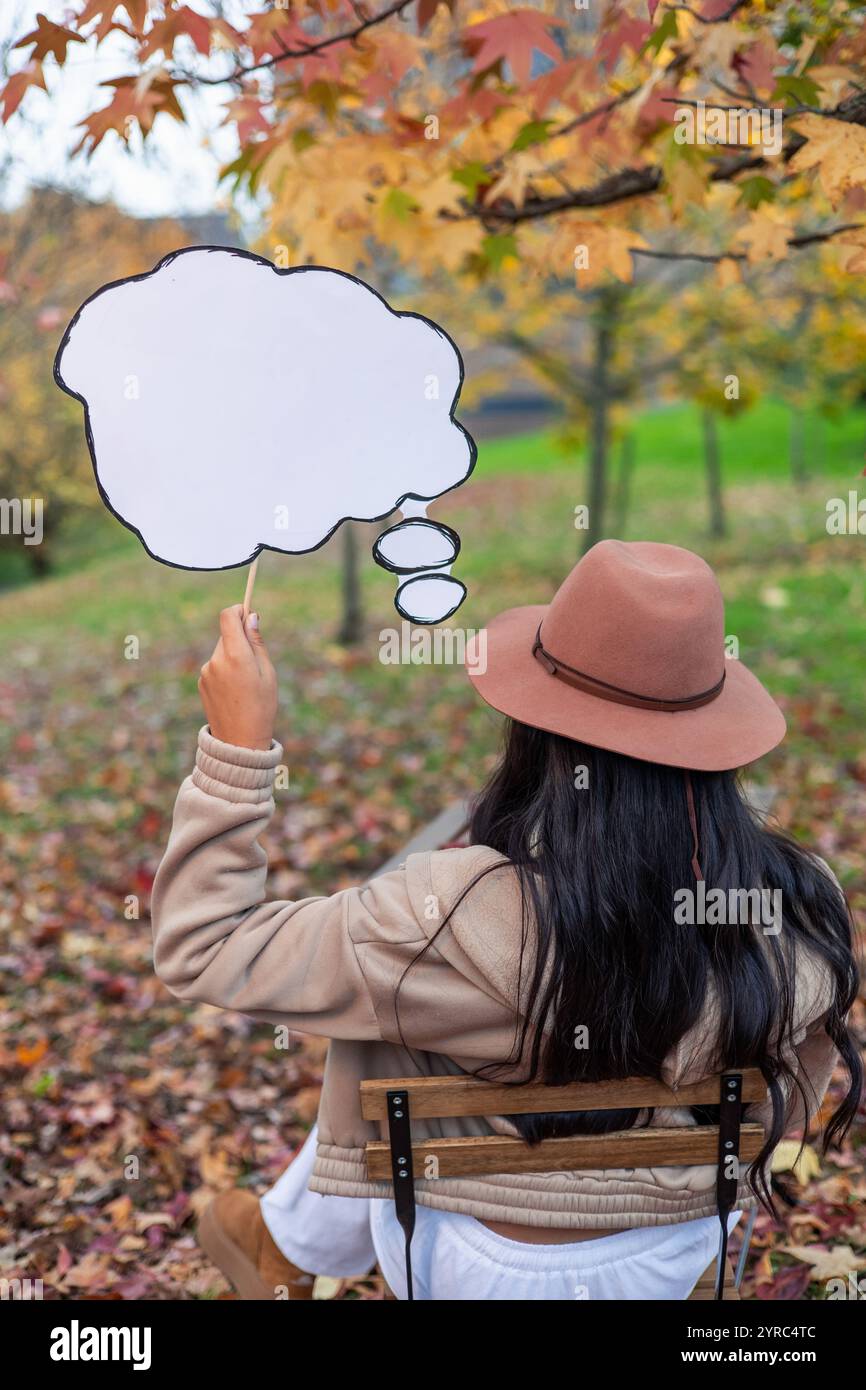 latin Woman wearing a hat is sitting on a chair in a park during autumn and holding an empty thought bubble, suggesting introspection, ideas, or imagi Stock Photo