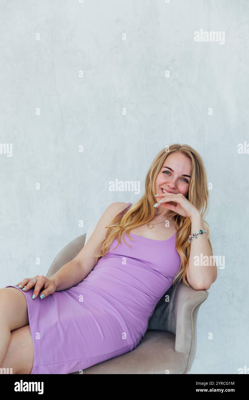 Beautiful Blonde Woman Smiling In Purple Dress Sitting On Chair Stock beautiful-blonde-woman-smiling-in-purple-dress-sitting-on-chair-stock