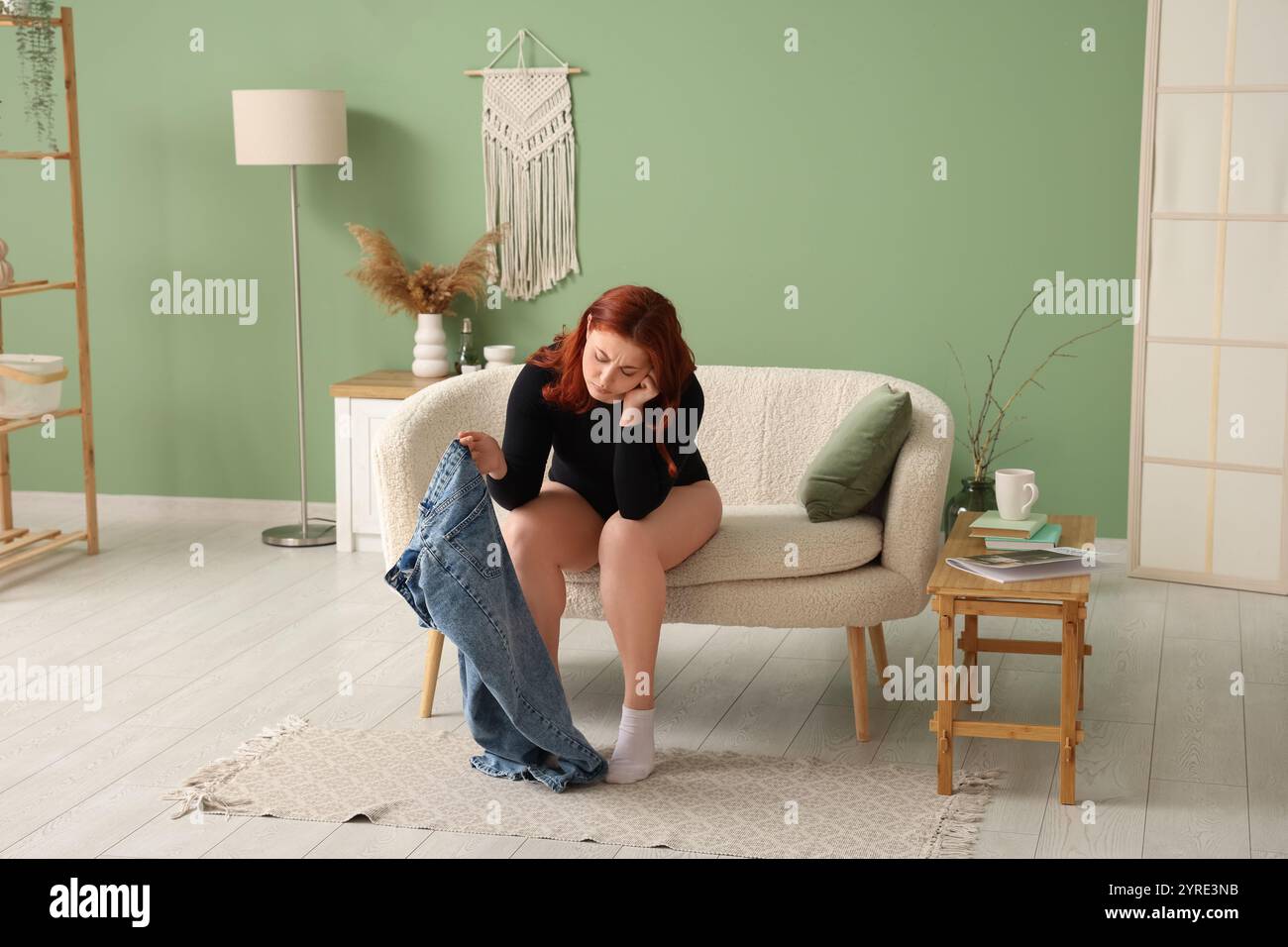 Upset young overweight woman with tight jeans sitting on sofa at home Stock Photo