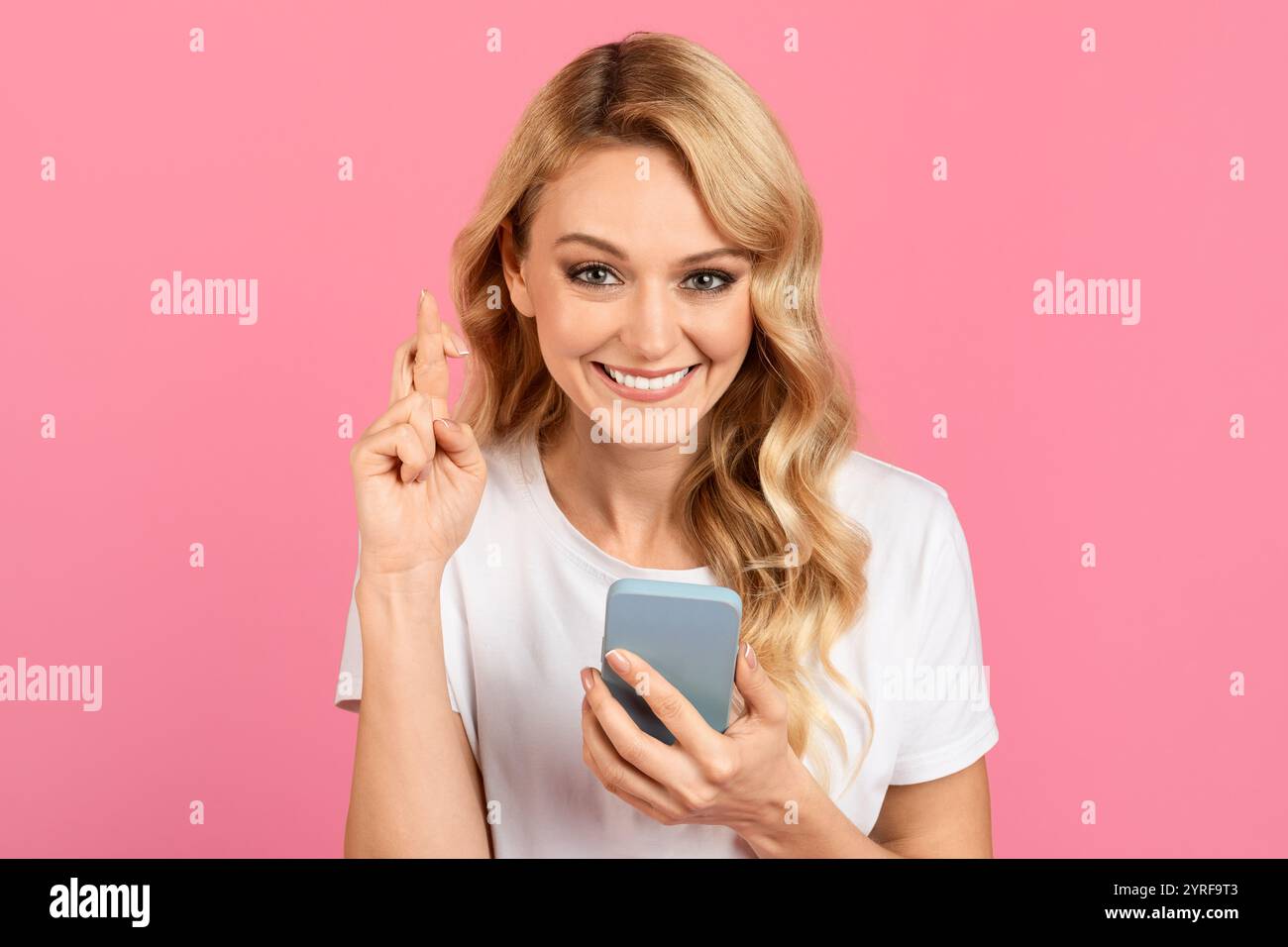 Woman expressing excitement while holding a smartphone against a pink background Stock Photo