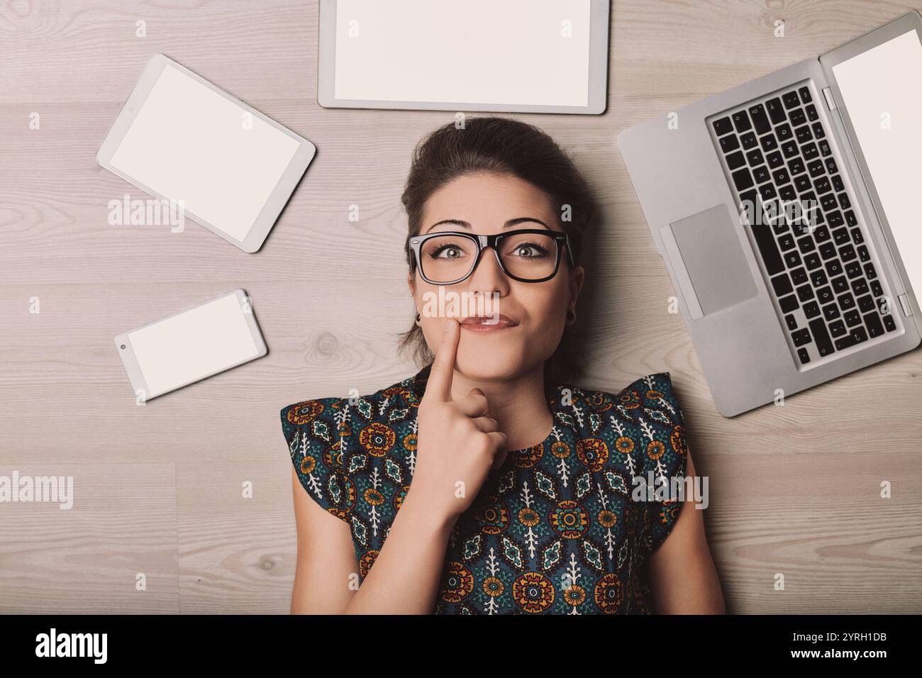 Young woman with eyeglasses lying on the floor surrounded by a laptop, tablet and smartphone, all with blank white screen, touching her chin with her Stock Photo