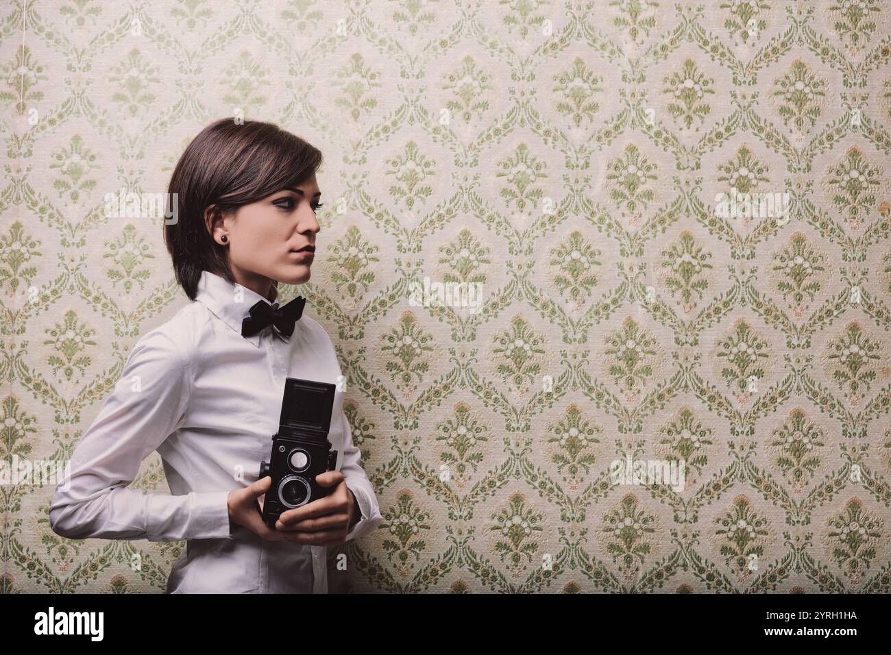 Young female photographer holding a vintage twin-lens reflex camera while posing against a patterned wallpaper, creating a nostalgic and artistic atmo Stock Photo