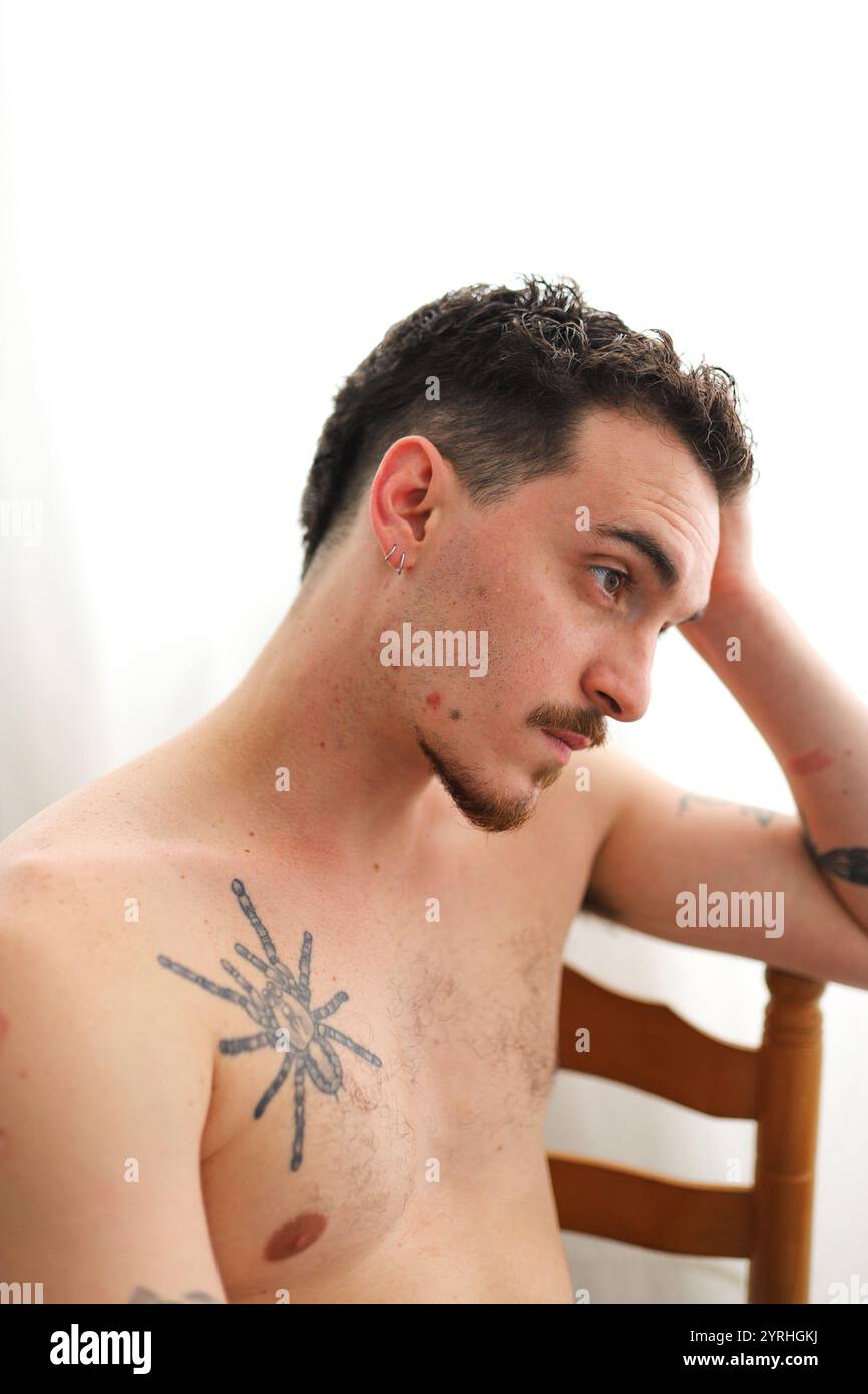 A young man with short hair and tattoos reflects while seated on a wooden chair. The minimalist white background highlights his contemplative expressi Stock Photo