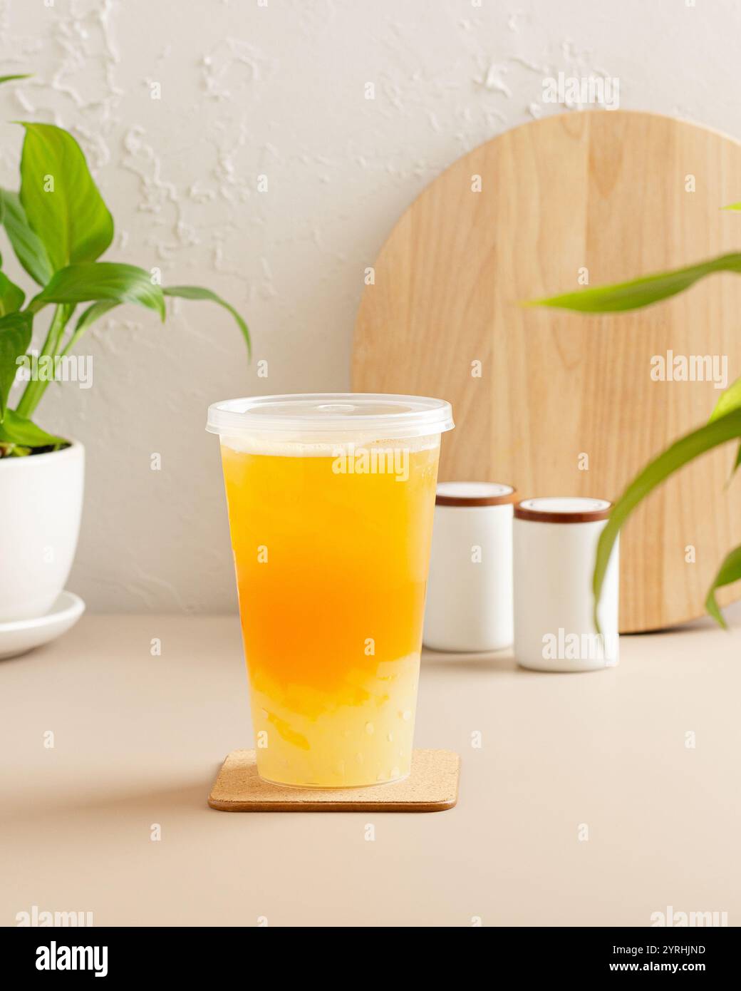 A tall cup of iced tea with a vibrant orange hue sits on a coaster, surrounded by green plants and simple white decor, creating a cozy and modern aest Stock Photo