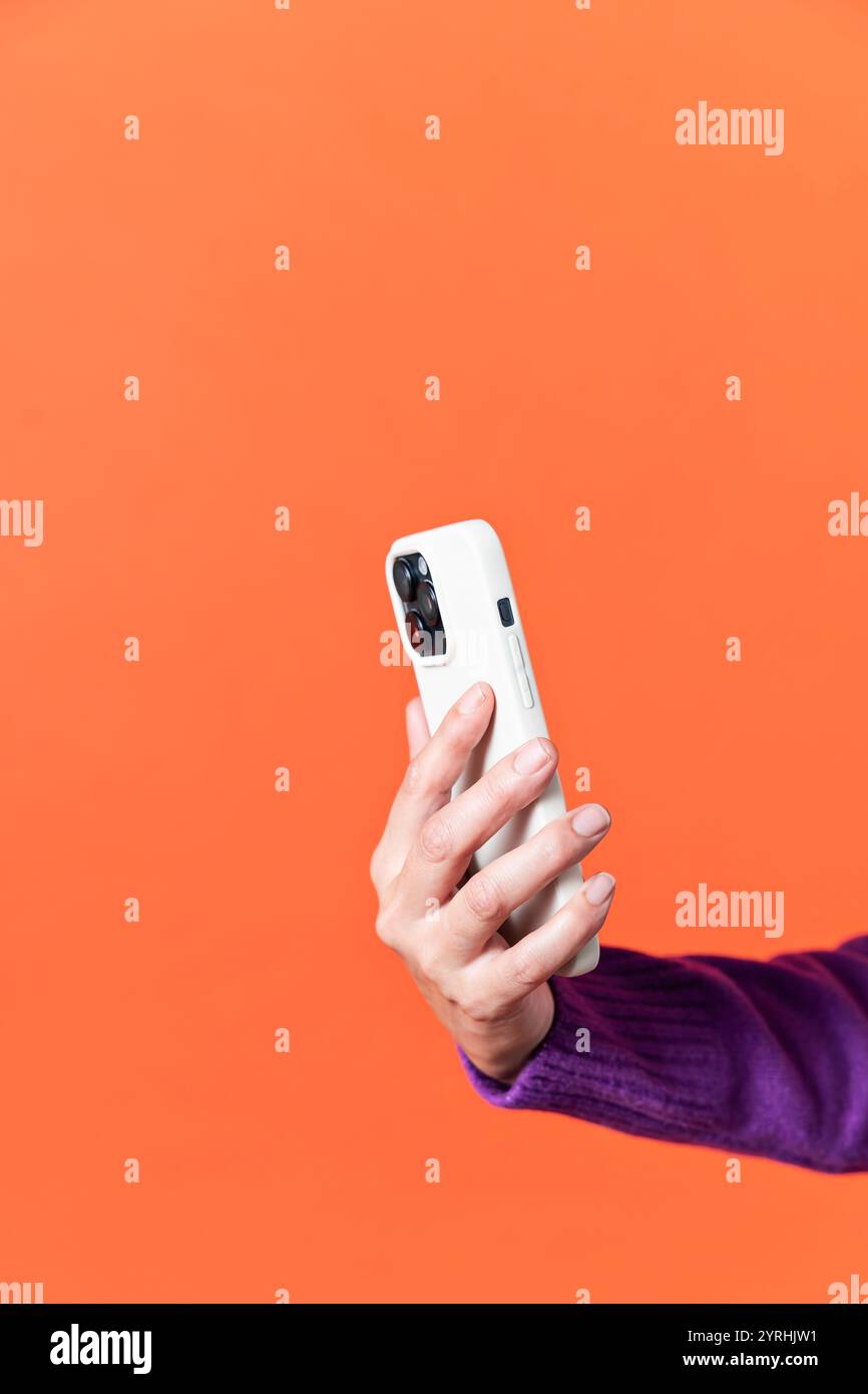 A close-up image of a female hand holding a white smartphone with a triple camera. The background is a vibrant, solid orange color, emphasizing modern Stock Photo
