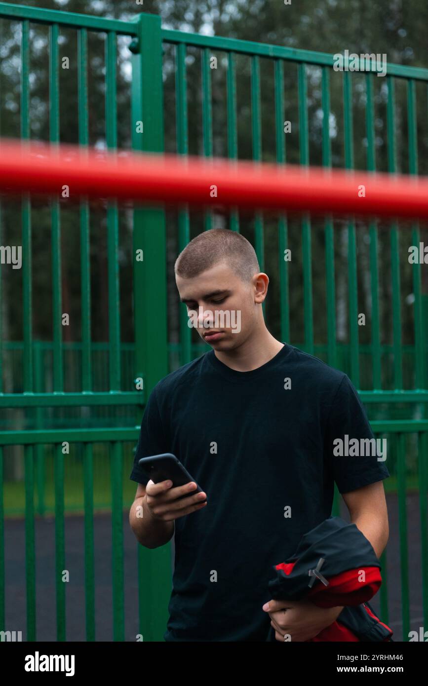 A young man stands in front of a green metal fence, looking intently at his smartphone. He holds a jacket and appears to be in a park or outdoor area, Stock Photo