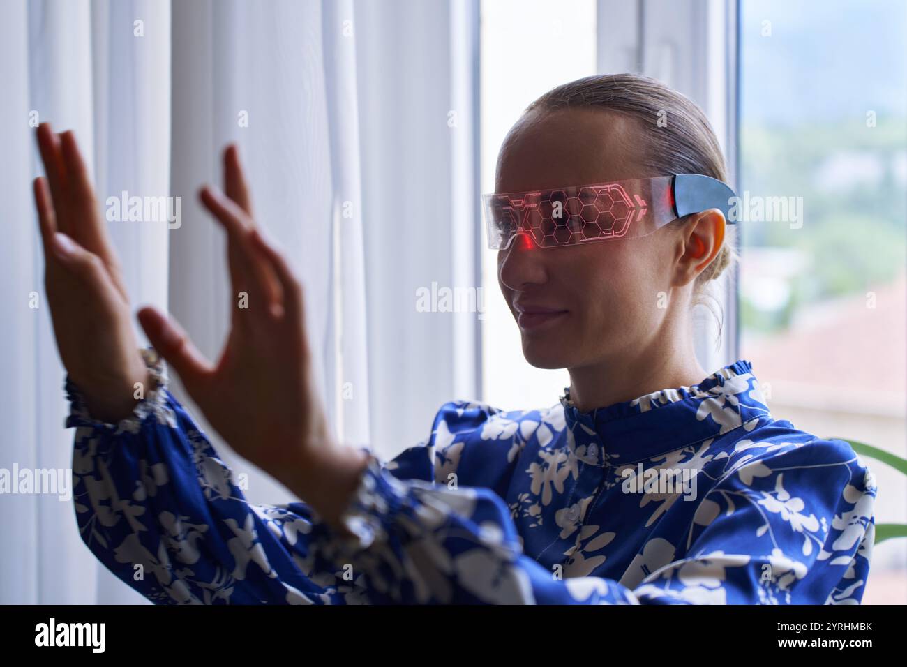 A woman in a blue floral blouse interacts with augmented reality using hi-tech, glowing glasses, standing by a window. The scene conveys innovation an Stock Photo