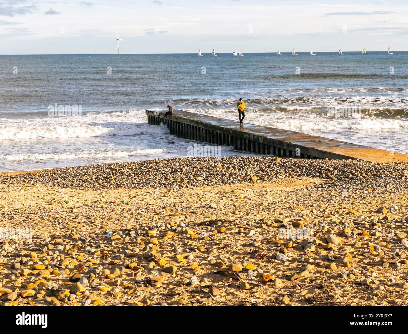 Pebble-strewn beach with a small pier extending into the sea, people standing on the pier enjoying the coastal view on a sunny day blyth south beach - Smartphone Captured Stock Image
