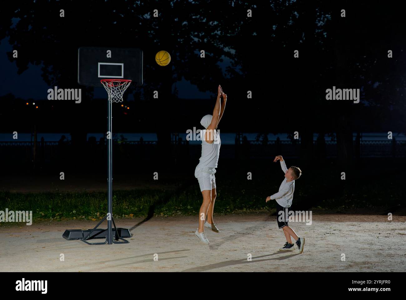 A father plays basketball with his young son at night in a park illuminated by a bright light Stock Photo