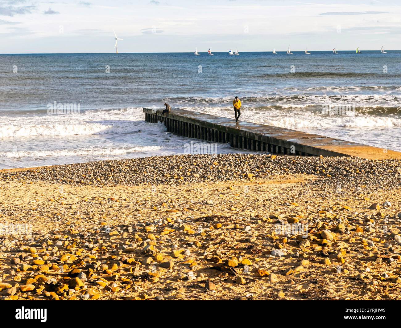 Pebble-strewn beach with a small pier extending into the sea, people standing on the pier enjoying the coastal view blyth south beach  northumberland - Smartphone Captured Stock Image