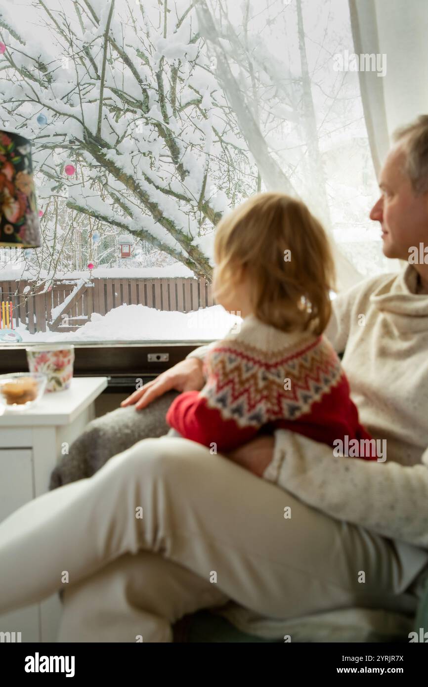 Middle age man and toddler sitting home in a chair by window with with scenic winter view of trees in snow. Bonding and quality family time together.  Stock Photo