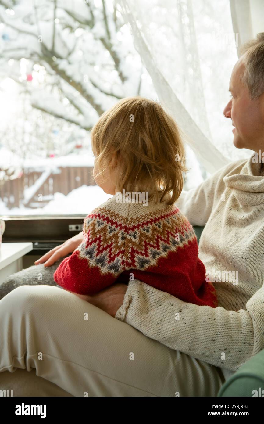 Middle age man and toddler sitting home in a chair by window with with scenic winter view of trees in snow. Bonding and quality family time together.  Stock Photo