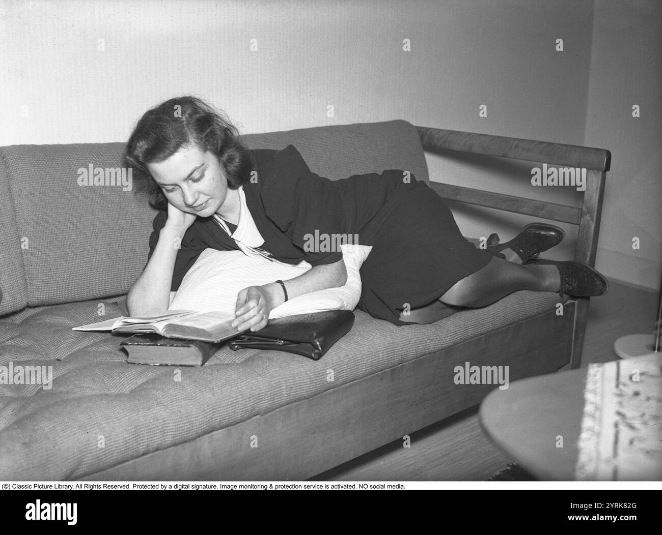 In the 1940s. A young woman at home reading from a book while lying ...