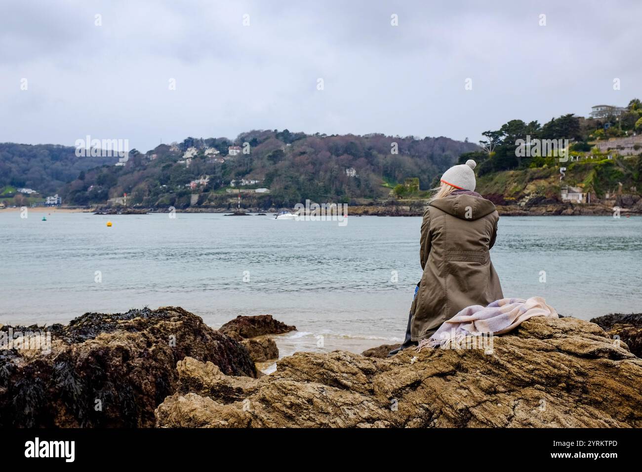 Blonde lady, in a bobble hat, sitting on a rock on Sunny Cove, East Portlemouth, watching the world go by with North and South sands in the background Stock Photo
