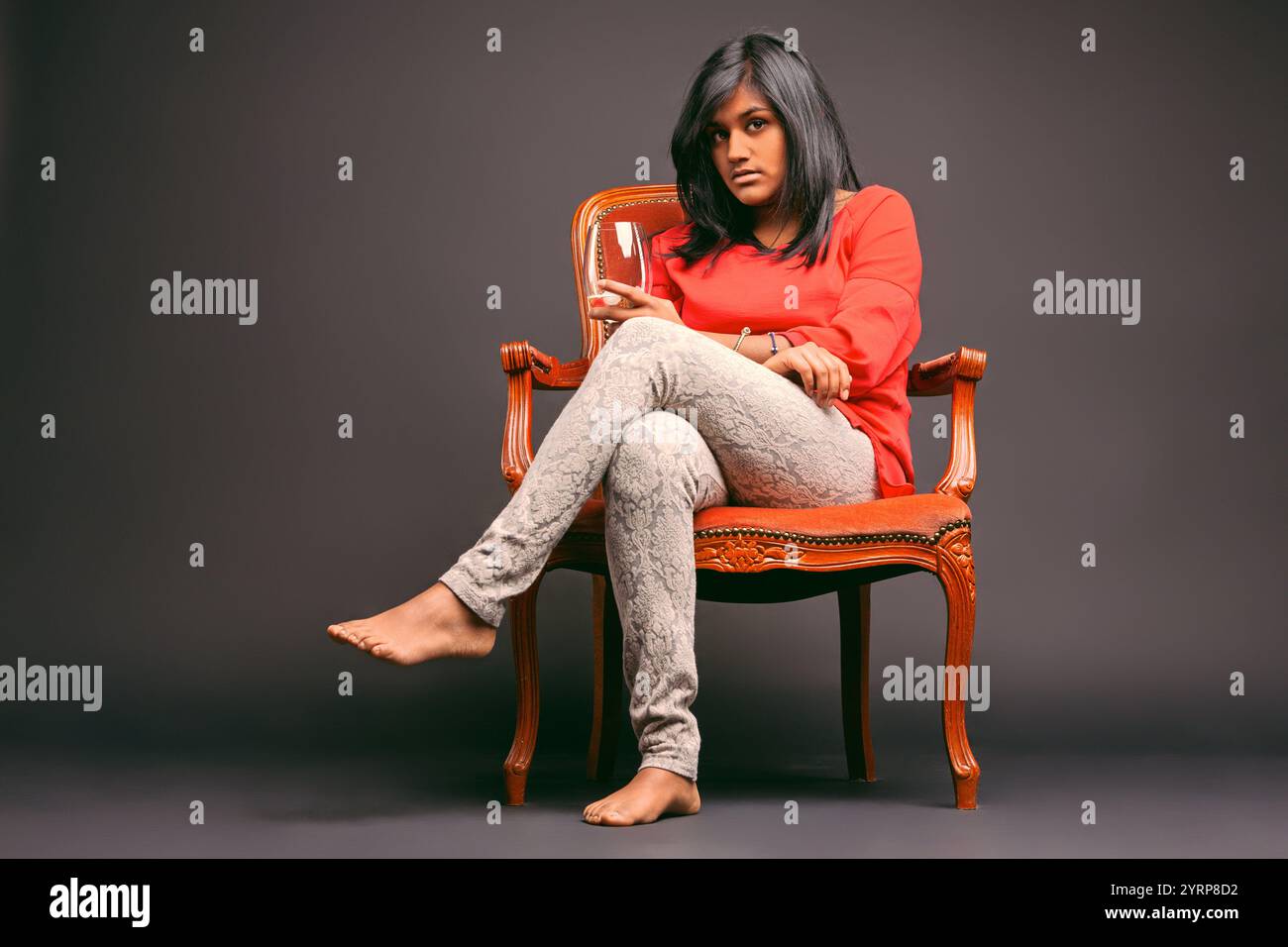 Young woman sitting barefoot on an elegant chair, holding a wine glass while gazing pensively at the camera against a soft gray backdrop, exuding an a Stock Photo