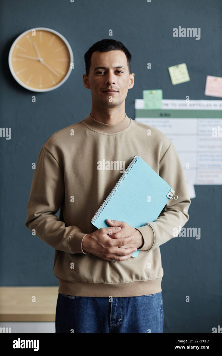 Vertical portrait shot of happy mixed raced male student posing with exercise book in hands while standing against blue wall in school classroom, copy space Stock Photo
