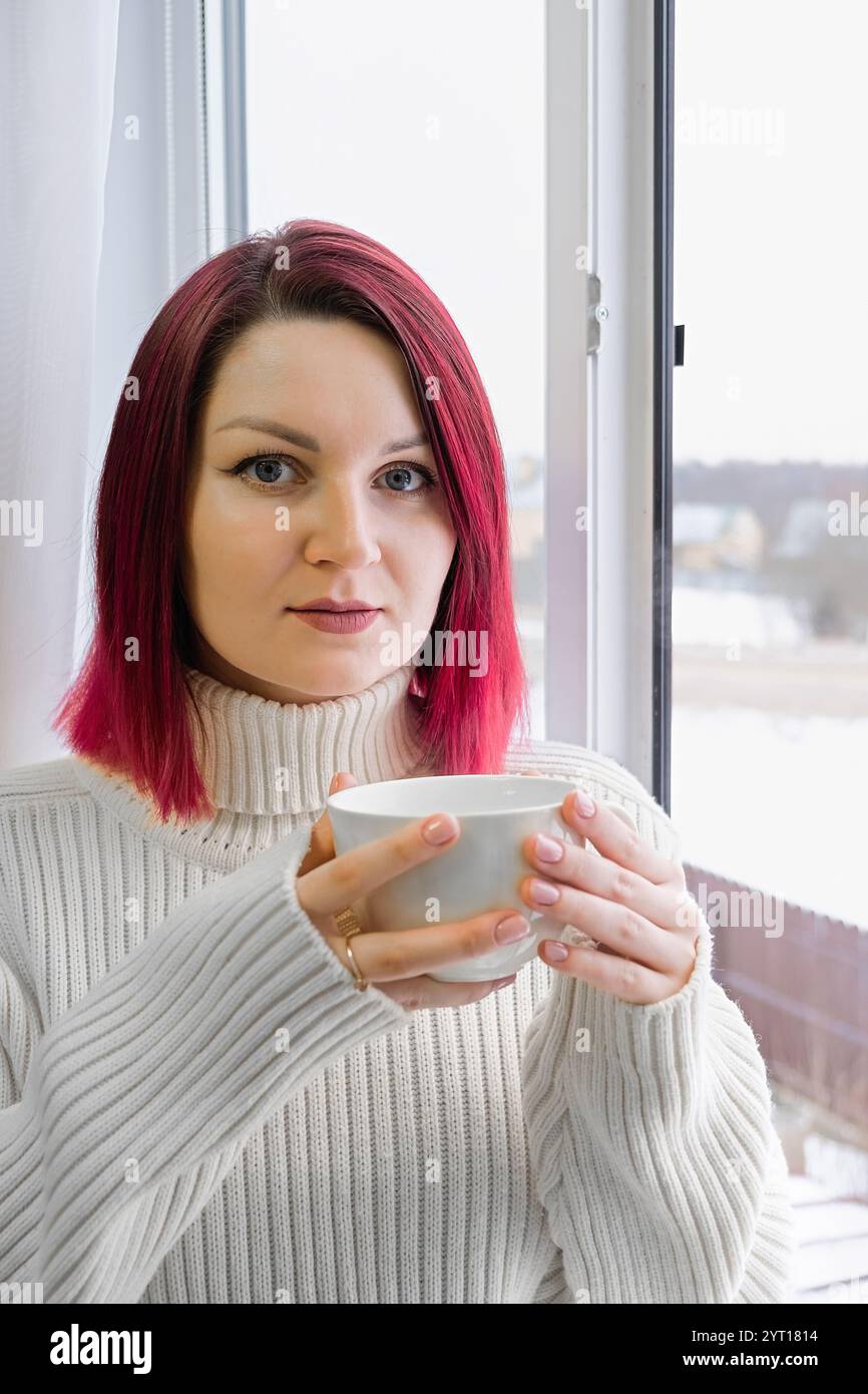 A woman with cherry red hair is holding a white cup of coffee. She is wearing a white sweater. Vertical crop. Closeup. Stock Photo