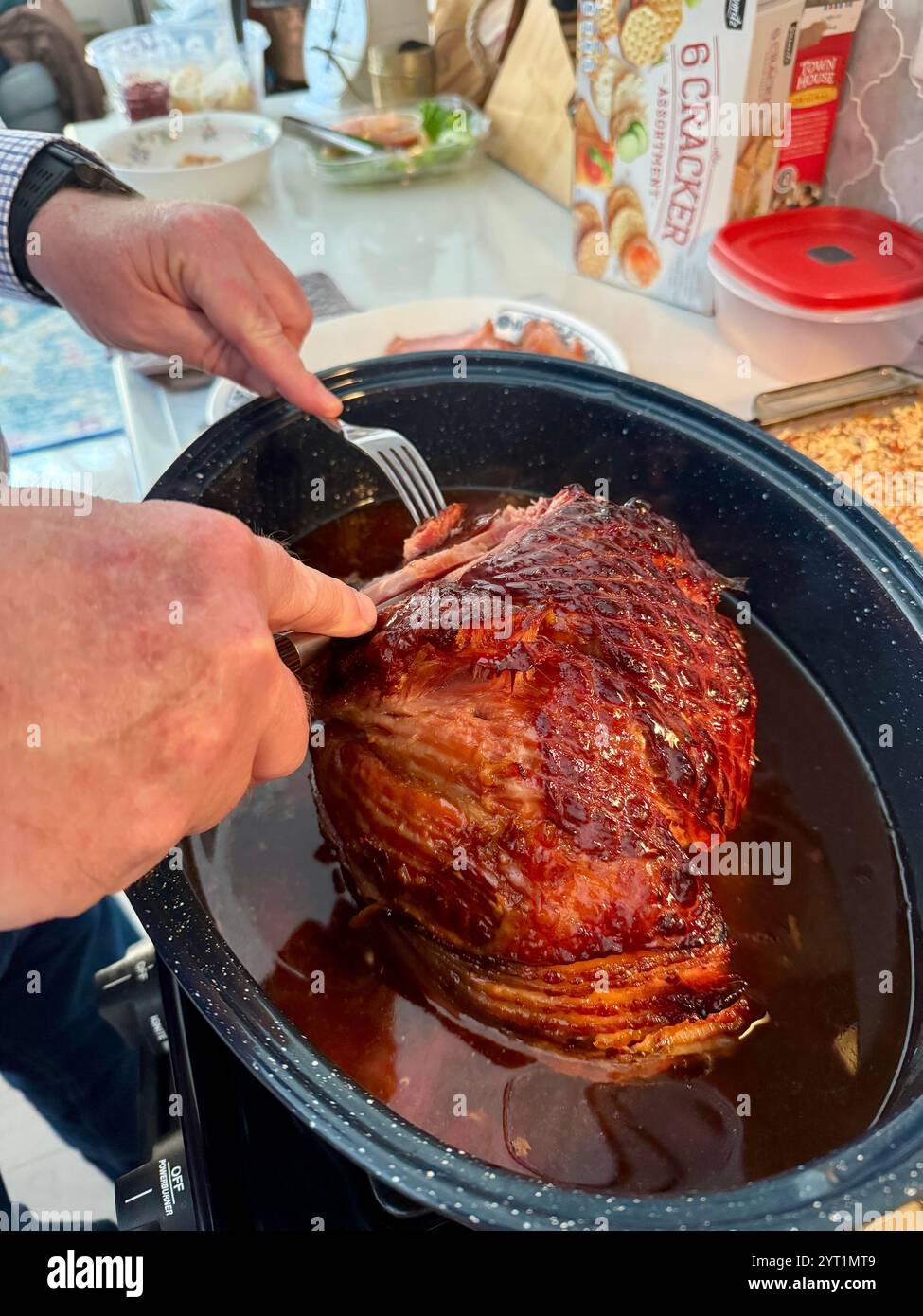 Holiday ham just out of the oven in roaster with glaze juices being sliced for serving with a man’s hands showing. USA - Smartphone Captured Stock Image
